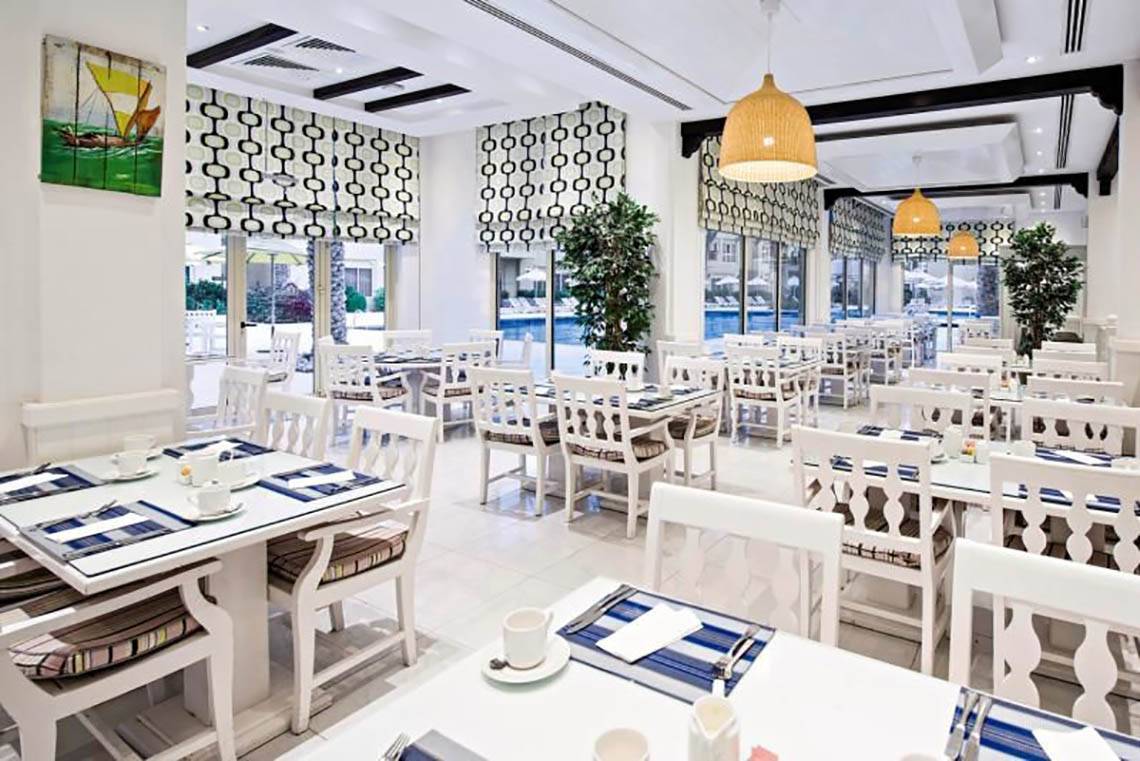 Innenansicht des Restaurants im Al Hamra Village Hotel mit weißen Tischen und Stühlen.