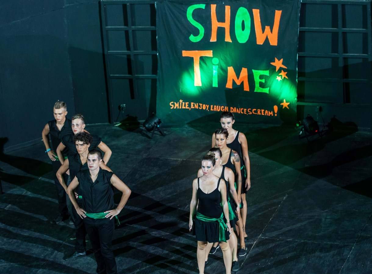 Tanzgruppe auf der Bühne unter einem 'Show Time'-Banner, das mit grünem Licht beleuchtet ist.