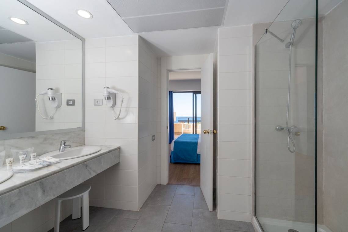 Badezimmer im Grand Teguise Playa mit Doppelwaschbecken, Spiegel, Föhnen und einer offenen Tür zum Schlafzimmer mit einem Bett.
