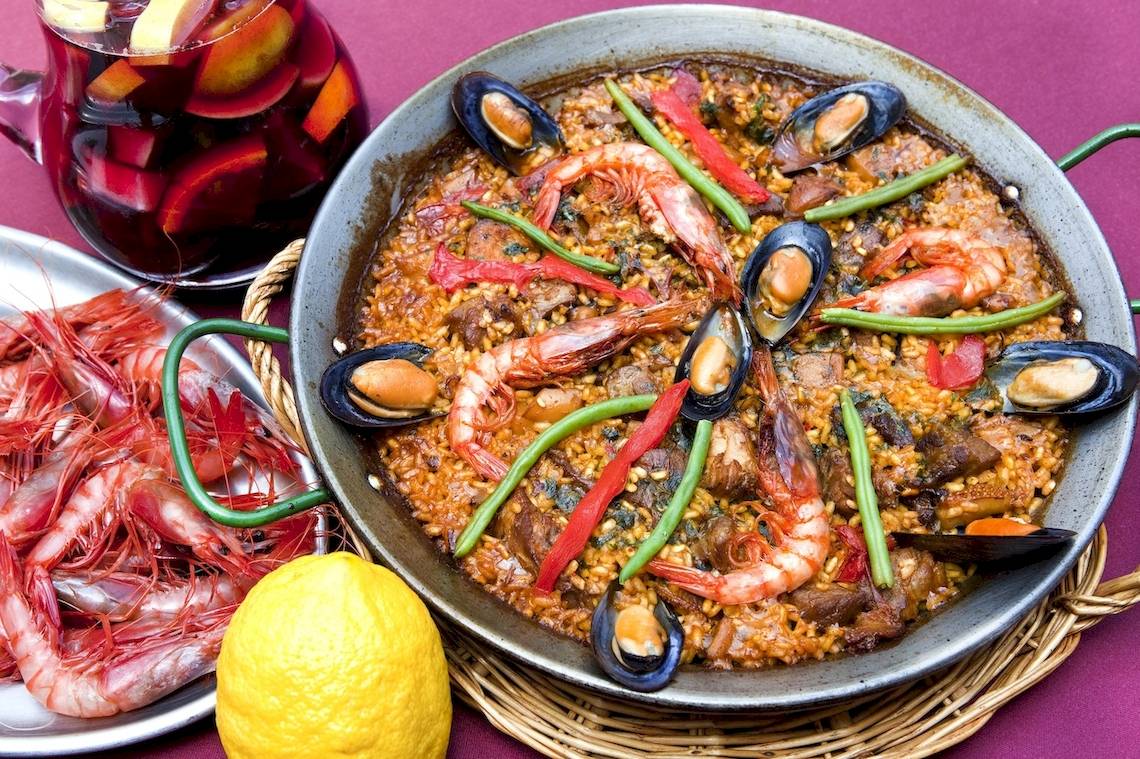 Eine Schüssel Paella mit Garnelen, Muscheln, Tomaten und Paprika auf einem Tisch neben einem Glas Sangria und einer Zitrone.