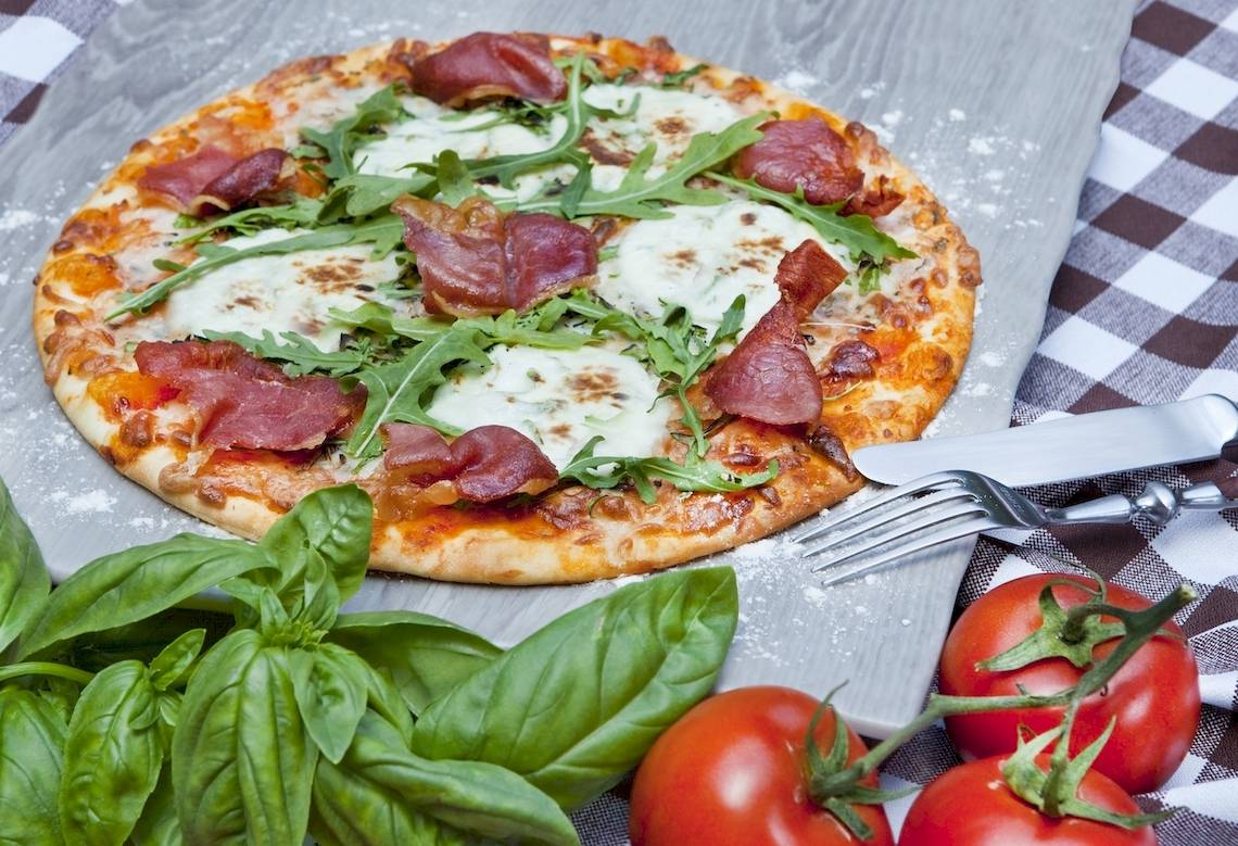 Eine Pizza mit Schinken, Rucola und Tomaten auf einem Marmorbrett.