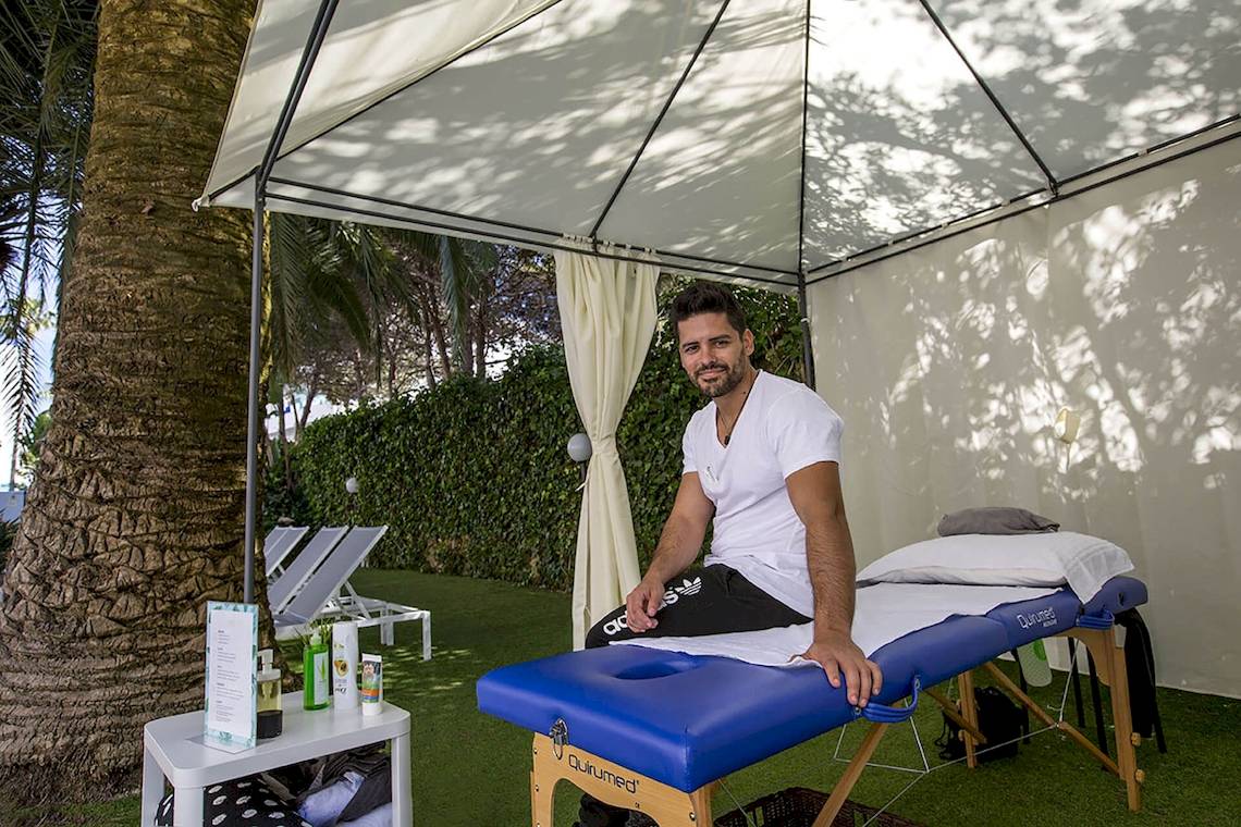 Ein Mann sitzt auf einem Massagestuhl unter einem Zelt im Freien, nahe einer Palme und mehreren Sonnenliegen, im Cosmopolitan Hotel.