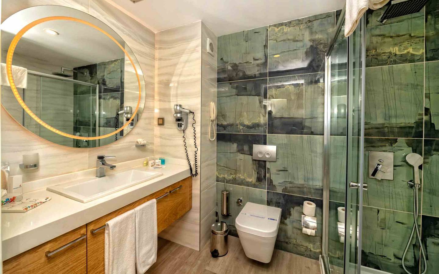 Badezimmer im Sidemarin Kirman Premium Hotel mit ovalem Spiegel, Waschbecken und Duschkabine.