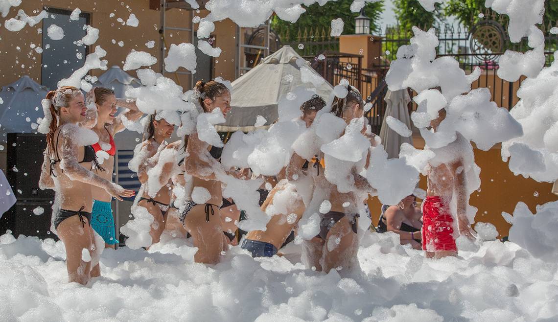 Mehrere Leute auf einem Seifenblasenfest in Badekleidung, umgeben von großen Seifenblasen.