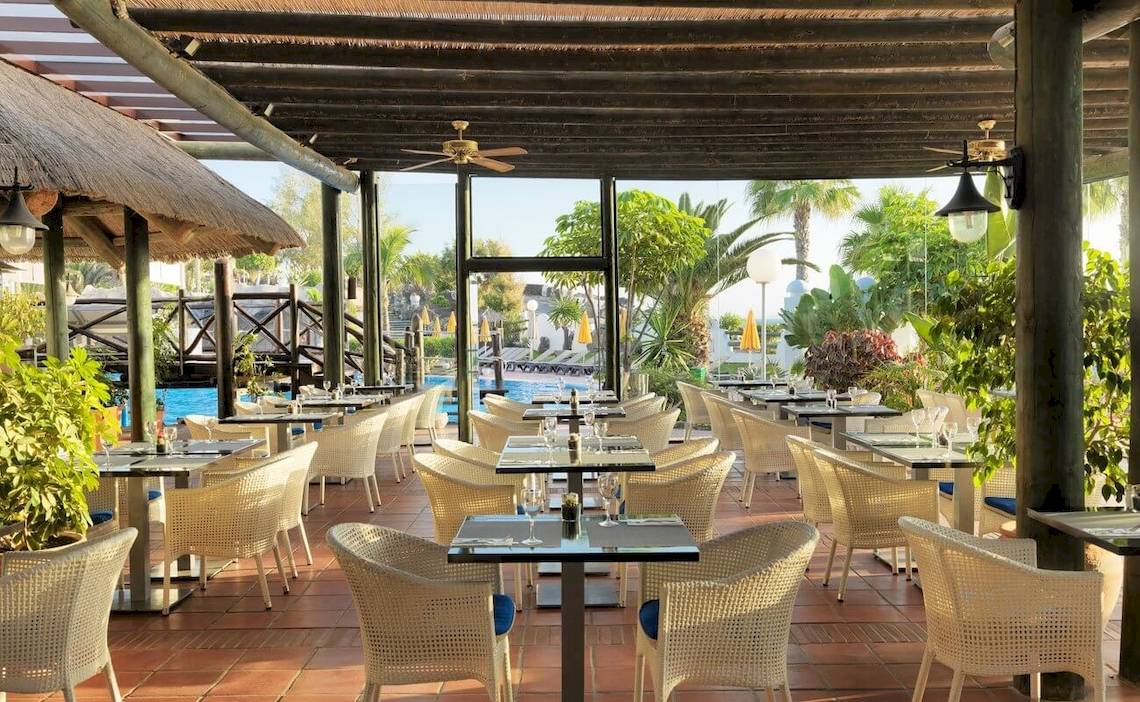 Innenbereich des Restaurants im H10 Timanfaya Palace mit Tischen und Stühlen, umgeben von Pflanzen und Blick auf den Pool.