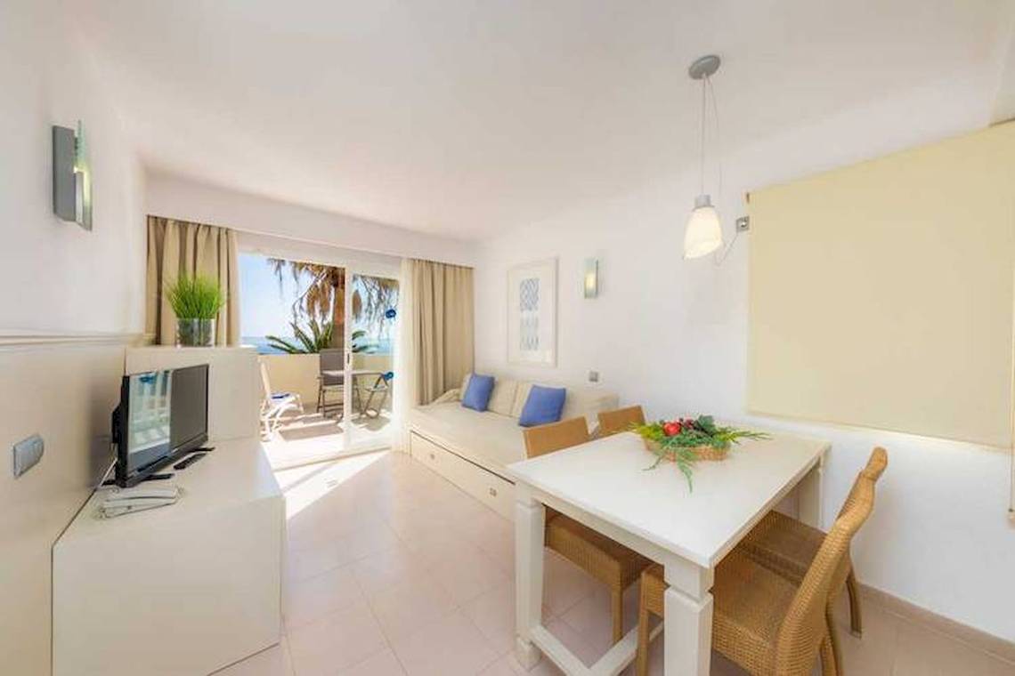 Wohnzimmer des Blau Punta Reina Hotels mit Tisch, Fernseher und offener Terrasse.