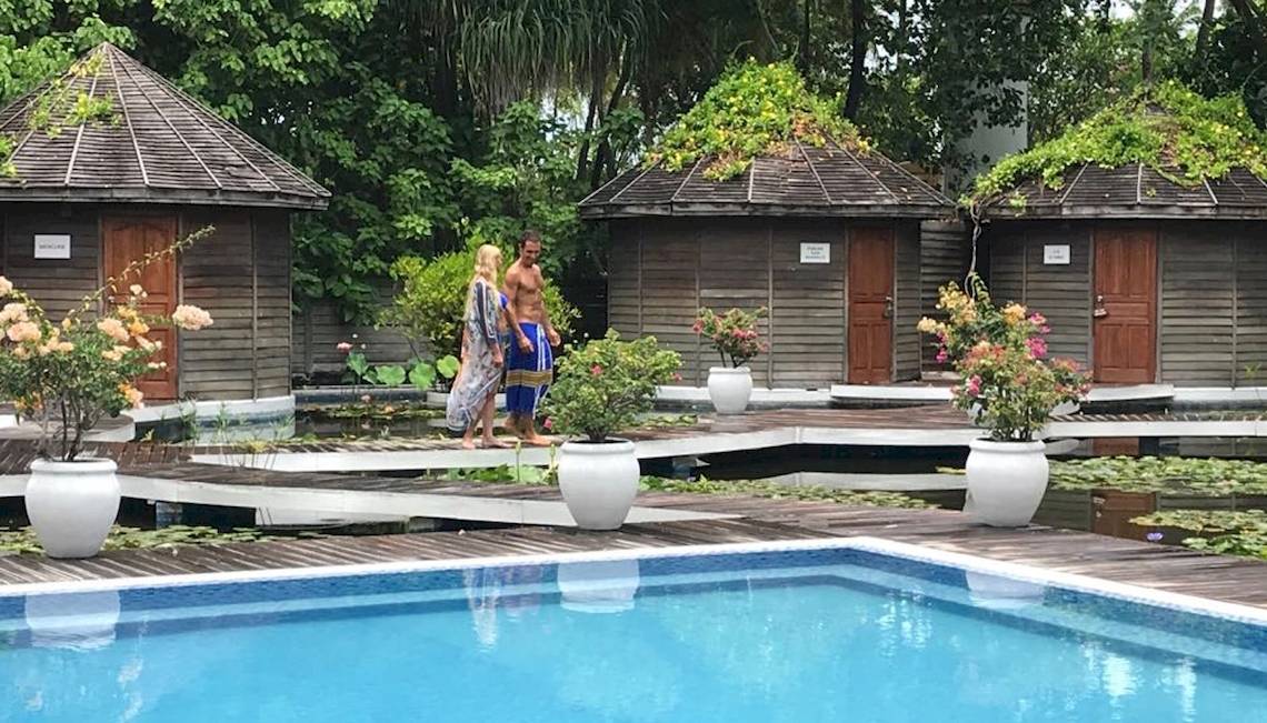 Ein Paar steht neben einem Schwimmbad im Freien, umgeben von blumengeschmückten Hütten und Pflanzen.