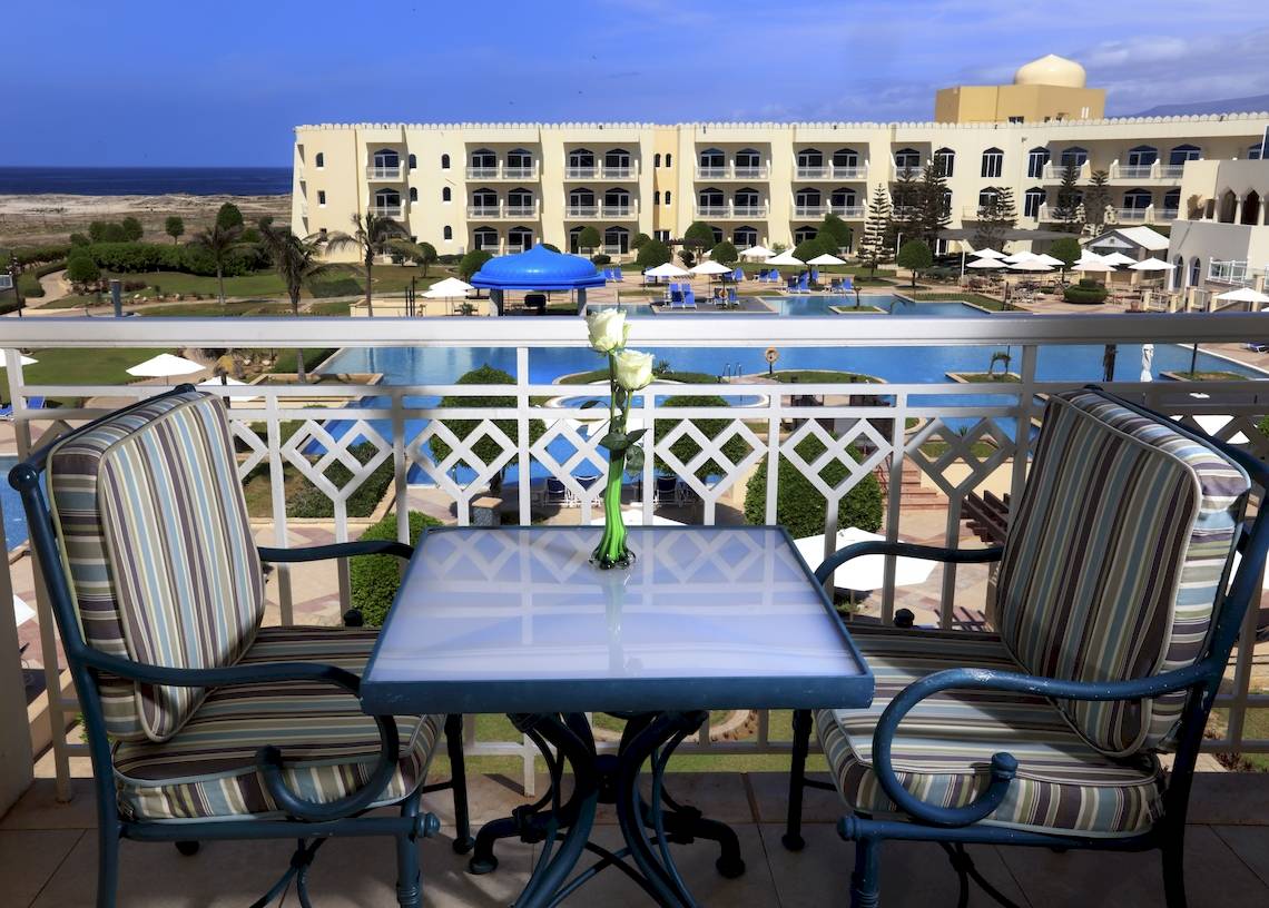 Ein Blick von einem Hotelbalkon mit Tisch und Blumen auf das Meer und das Wyndham Garden Salalah Mirbat Hotel.