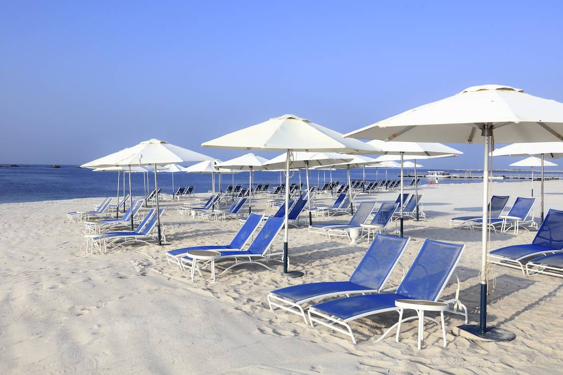 Strandszene mit vielen weißen Sonnenschirmen und blauen Strandliegen im Sand