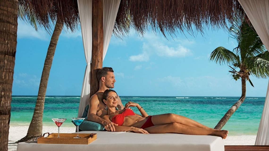 Paar im roten Bikini und Anzug entspannt auf einem Strandbett unter einem Sonnenschirm, mit Blick auf das Meer, Palmen und Cocktails.