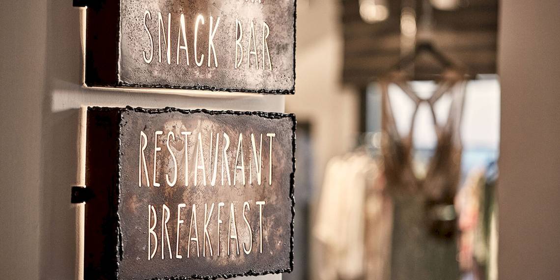 Weiß beleuchtete Schilder an einer Wand im Myconian Avaton Resort, die Snack Bar und Restaurant Breakfast anzeigen.