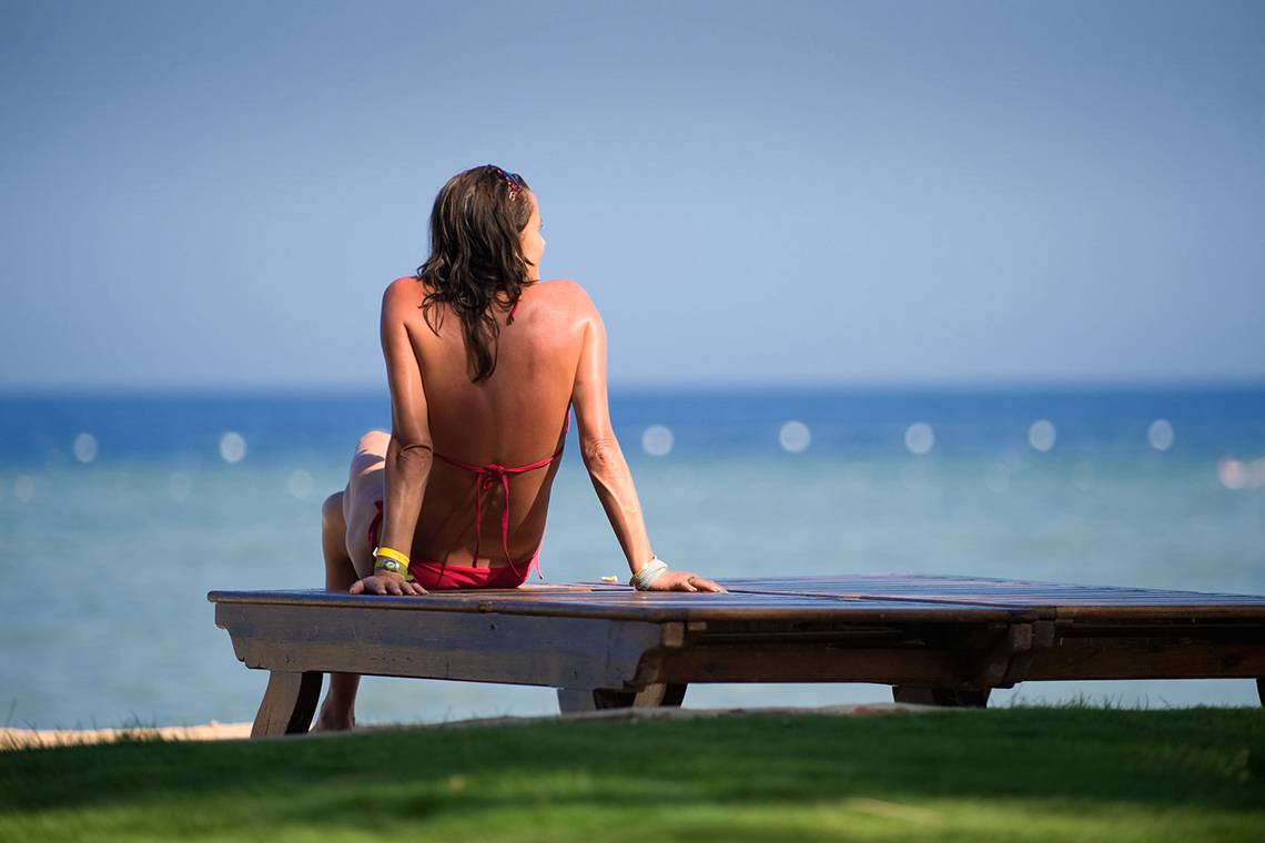Frau im roten Bikini sitzt auf einer Bank am Strand und blickt auf das Meer.