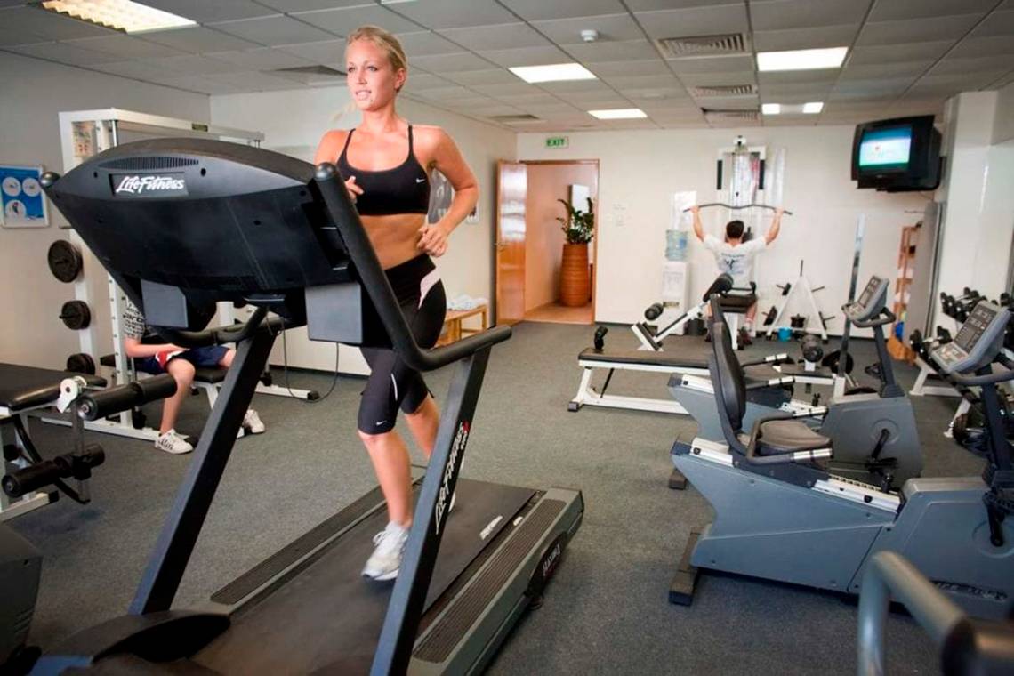 Frau trainiert im Fitnessstudio auf einem Laufband, andere Geräte im Hintergrund.