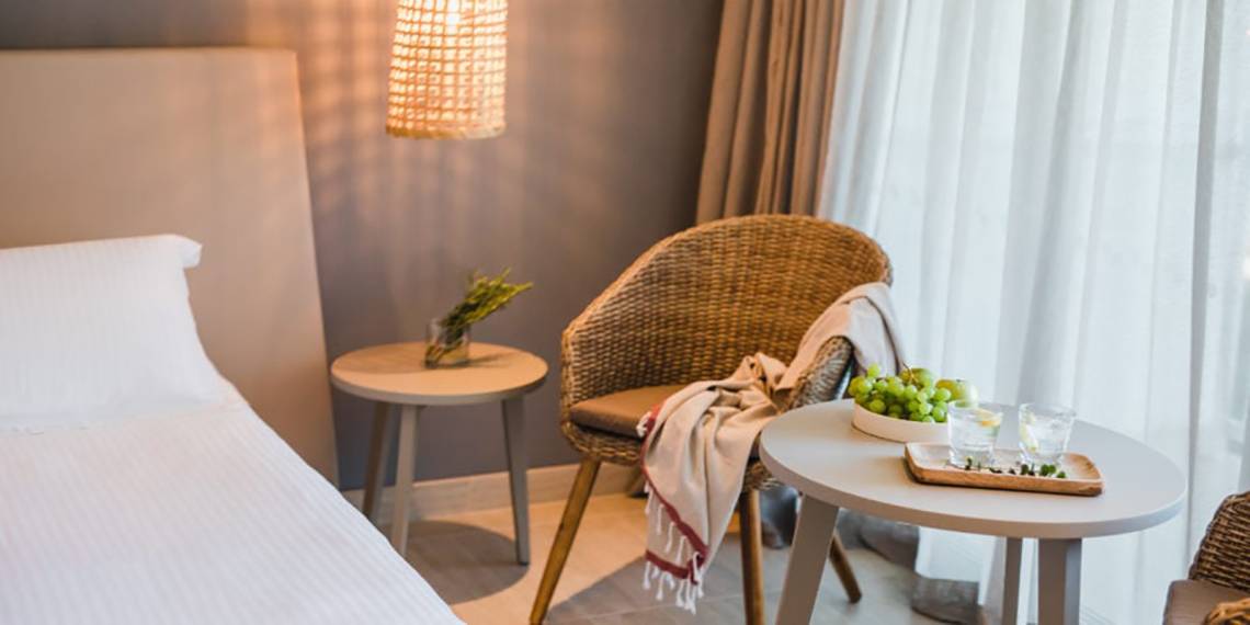 Innenansicht eines Zimmers im Nissi Beach Resort mit Bett, zwei Nachttischen, einem Korbstuhl und einem Tisch mit Früchten.