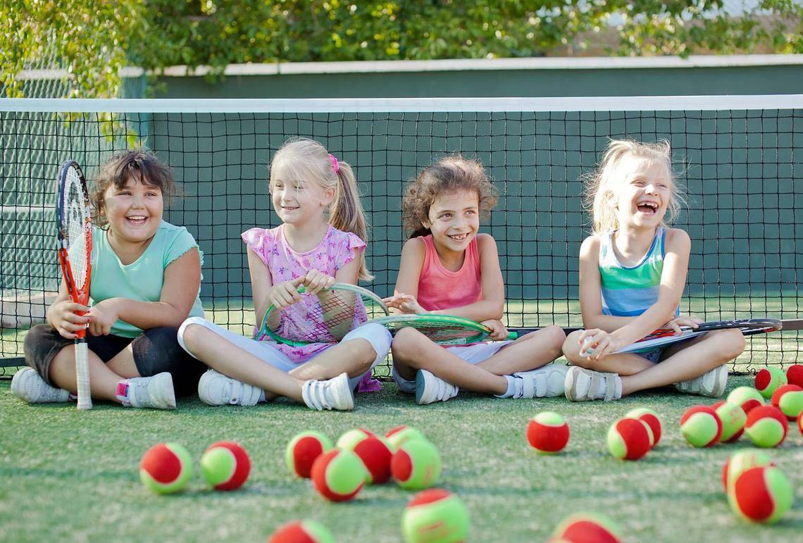 Vier Maedchen sitzen auf einem Tennisplatz, lachen und halten Tennisschlaeger in der Hand, mit Tennisbaellen um sie herum.
