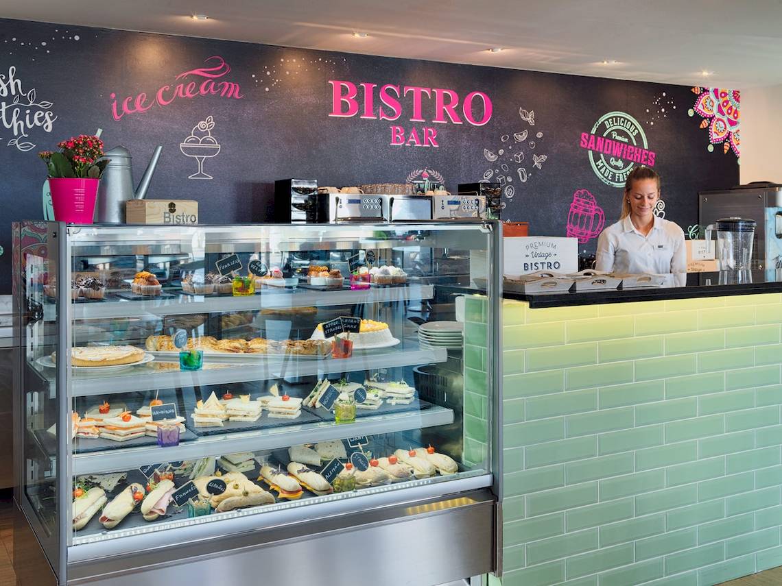 Innenansicht der Bistro Bar im H10 Rubicon Palace mit einer Wandtafel, die 'Bistro Bar' und 'ice cream' beschriftet ist, einer Verkäuferin und einer Vitrine voller Kuchen und Sandwiches.