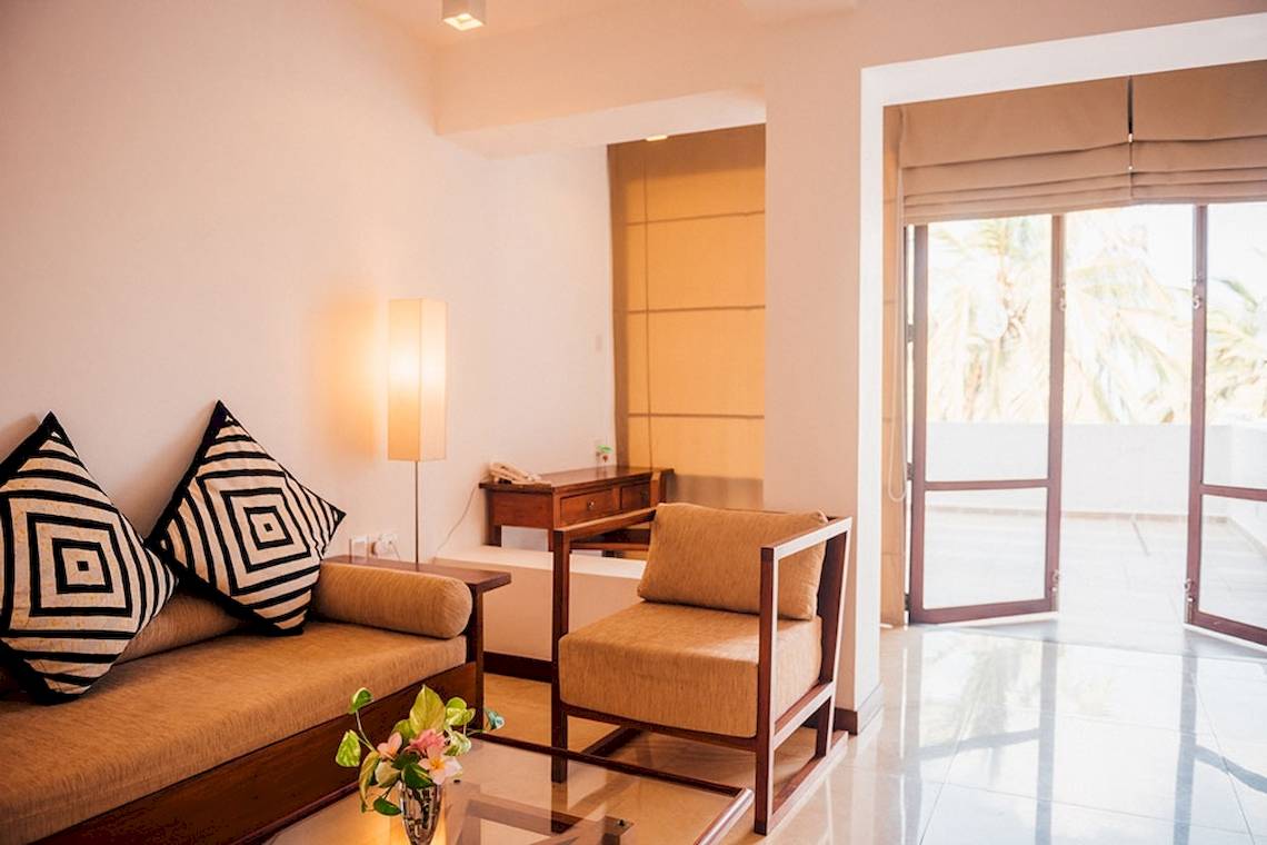 Wohnzimmer im Goldi Sands Hotel mit beigefarbener Couch, Kissen, Lampe, Tisch und Fenstern.