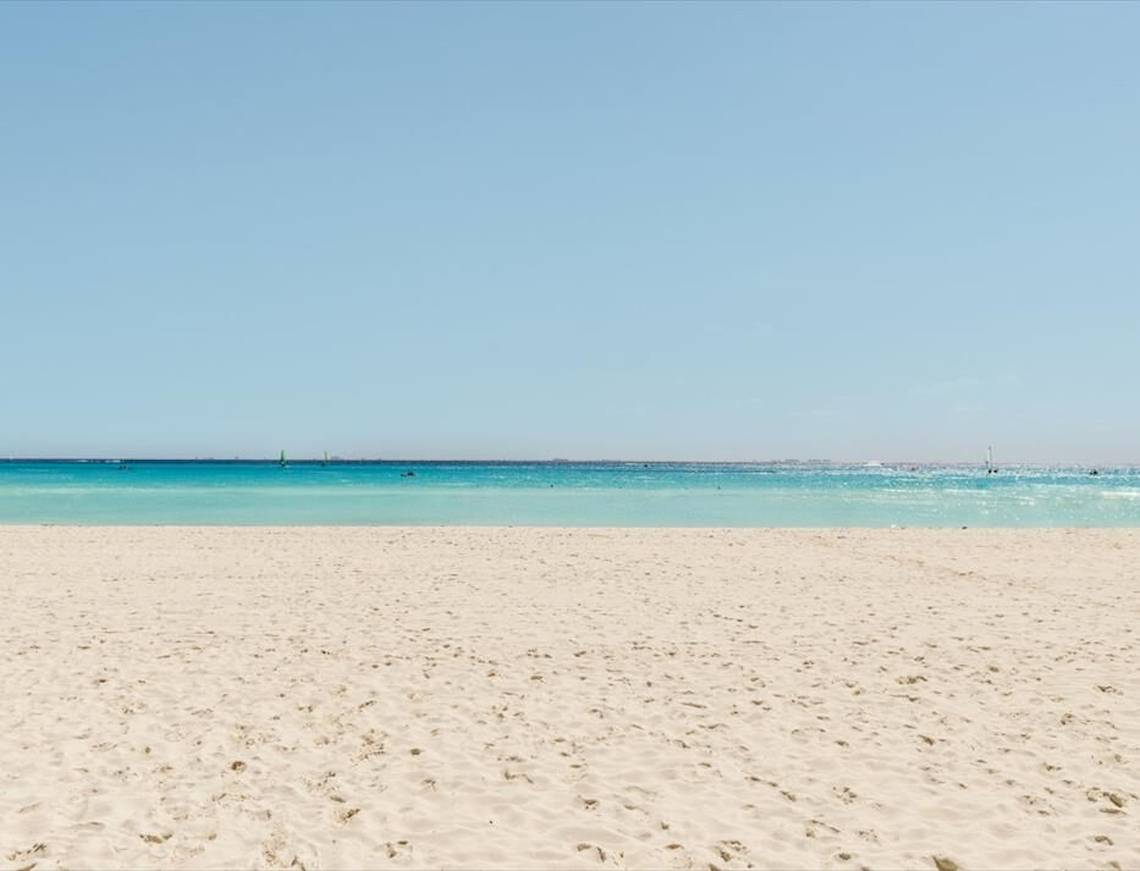 Foto von einem weißen Sandstrand und türkisfarbenem Wasser, mit einem blauen Himmel.