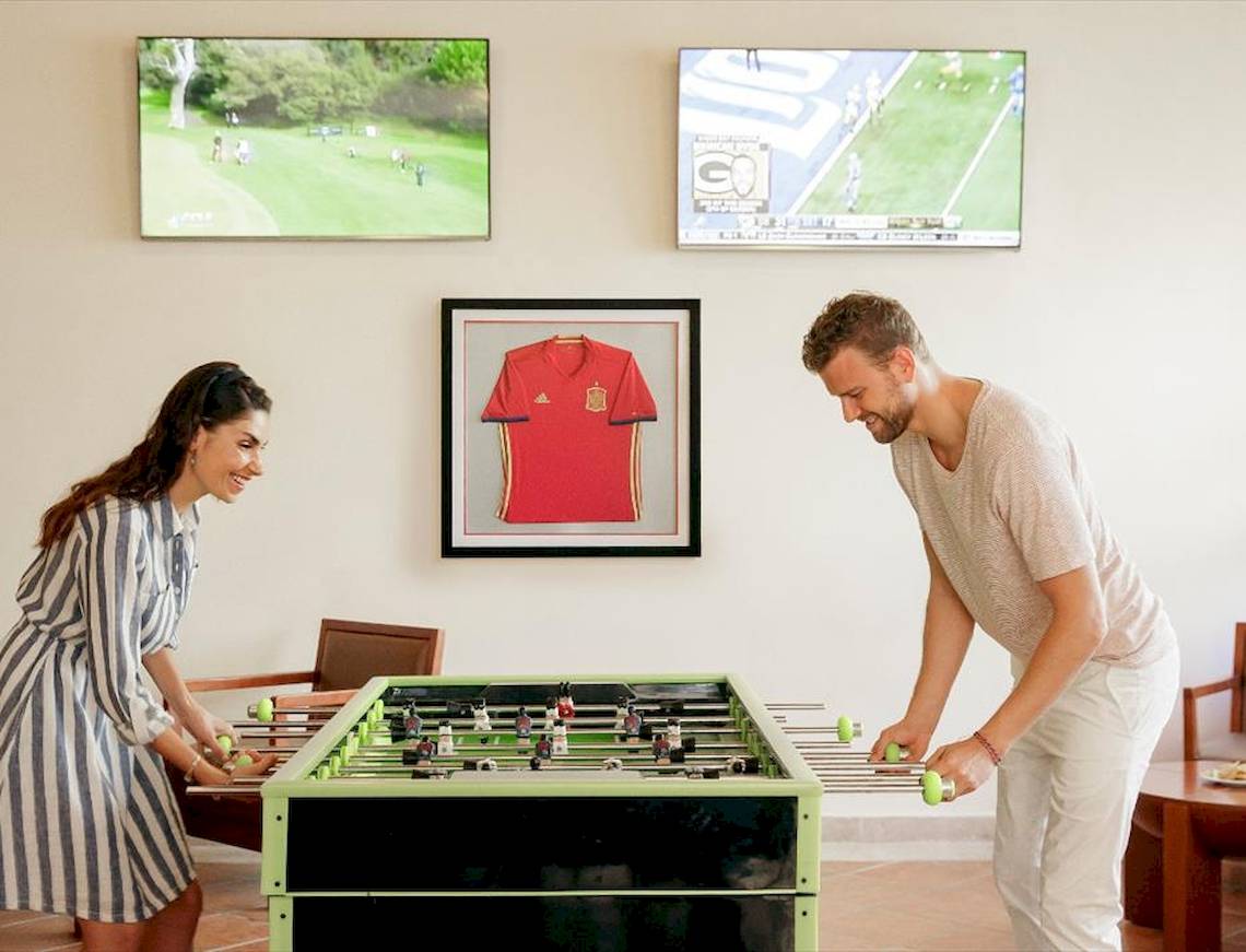 Gäste im Allegro Playacar Hotel spielen Tischfußball vor einem Fußballtrikot und Fernsehern an der Wand.