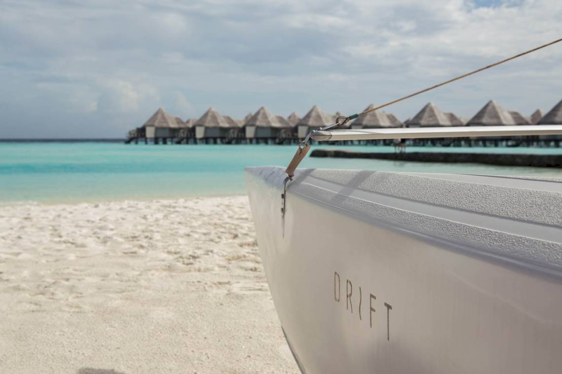 Drift Thelu Veliga Retreat Boot am Strand mit Blick auf Wasser und traditionelle Hütten.