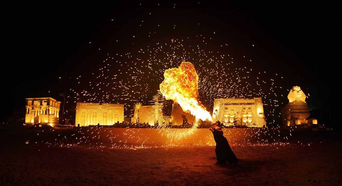 Eine Person spuckt Feuer in der Nacht vor alten Gebäuden und einer Sphinx-Statue.
