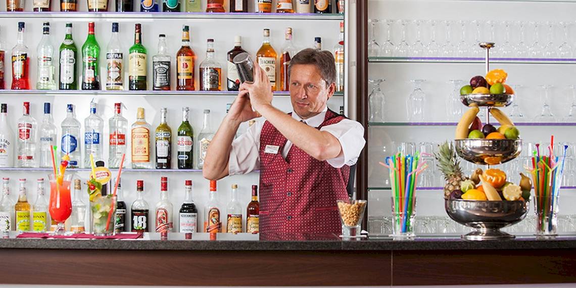 Ein Barkeeper im roten Jackett des Zora Hotels rührt einen Cocktail in einem Glas.