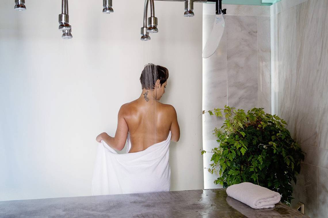 Frau im Kontokali Bay Resort & Spa steht unter einer Dusche und hält einen weißen Bademantel.