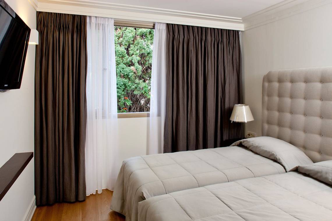 Schlafzimmer mit zwei Betten, Fenstern, Vorhängen, Fernseher und Lampe.