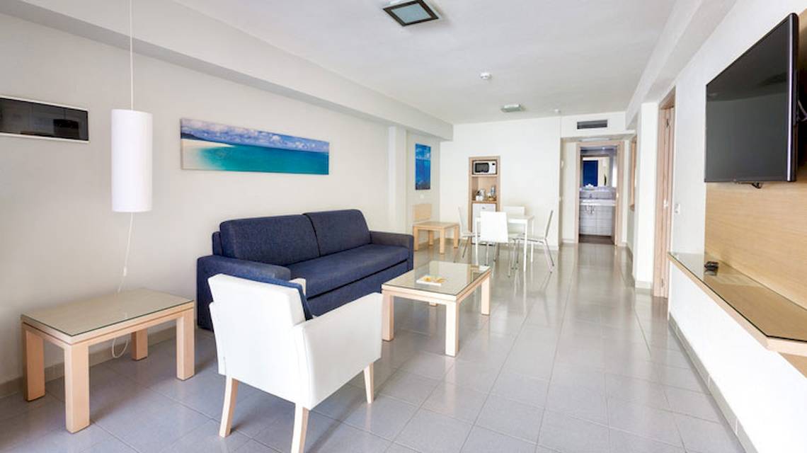 Inneneinrichtung des Tagoro Family & Fun Costa Adeje mit einem blauen Sofa, weißem Stuhl, Glastisch und Fernseher an der Wand.