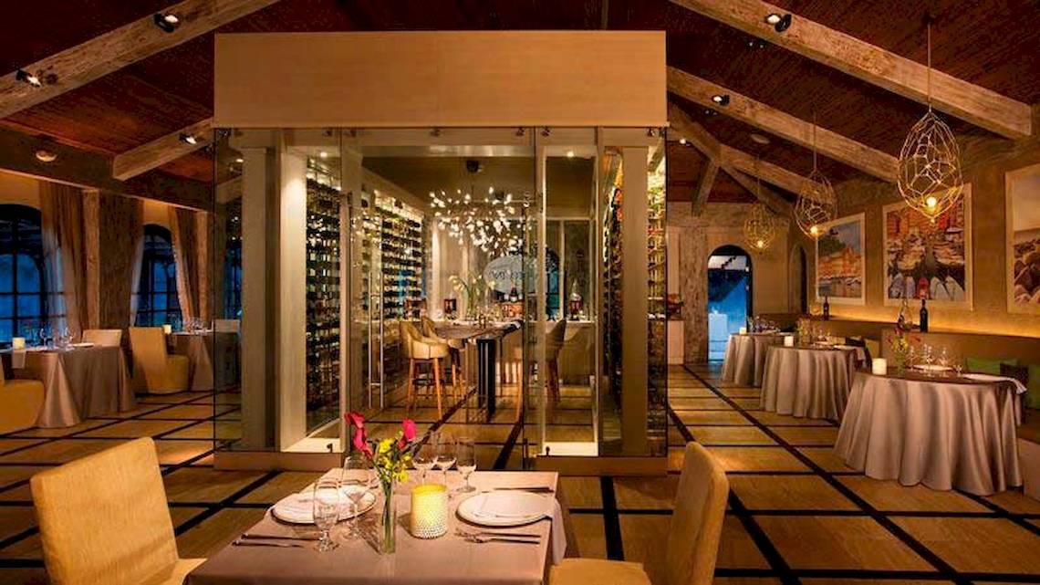 Innenbereich des Restaurants im Secrets Papagayo Costa Rica mit mehreren Tischen und Stühlen, einer großen Glaswand, Weinregal und dekorativen Lampen.