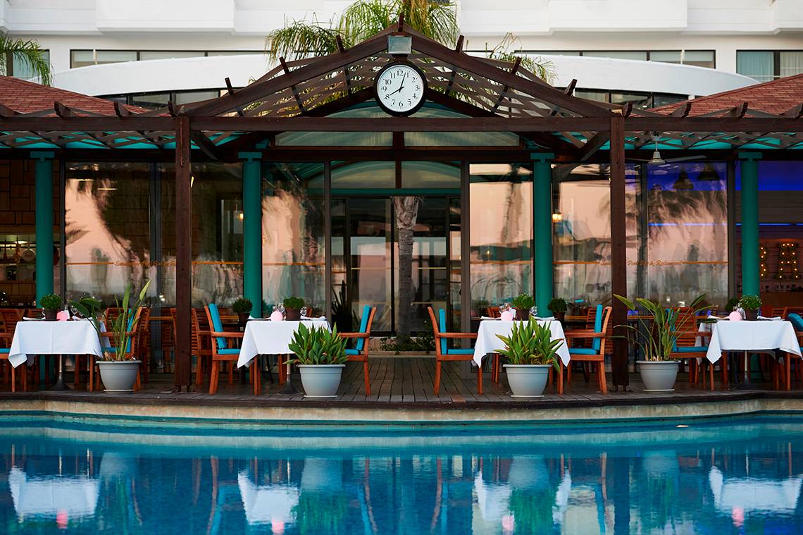 Pool-Lounge-Bereich im Hotel Louis Ledra Beach mit Tischen und Stühlen, Pflanzen und einer Uhr.