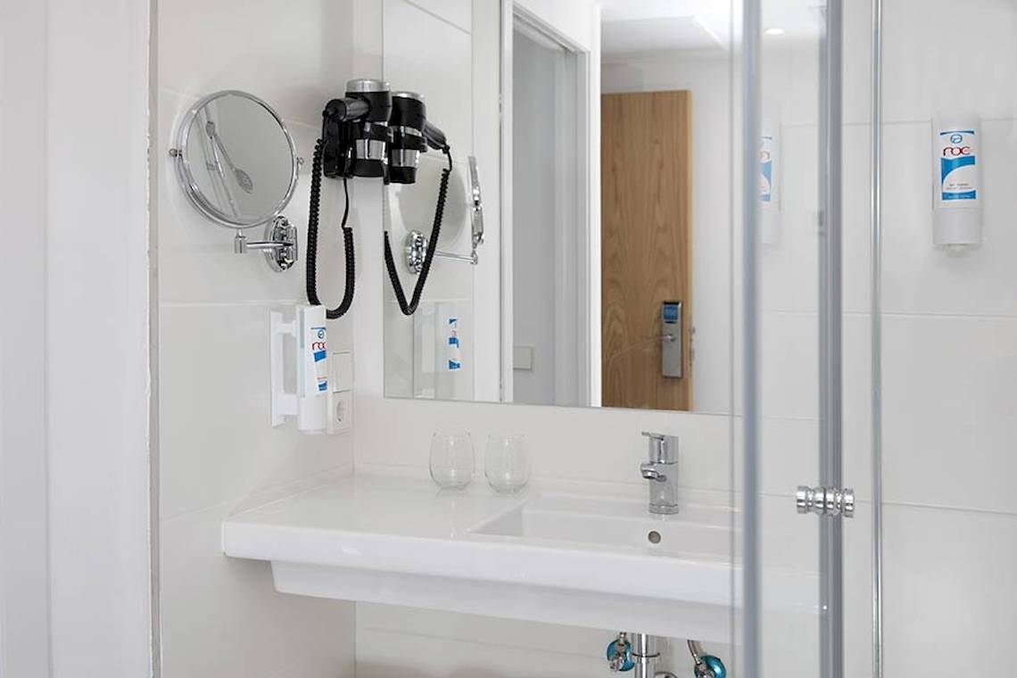 Badezimmer im Alua Leo Hotel mit Föhn, Spiegel, Waschbecken und Glastüren.