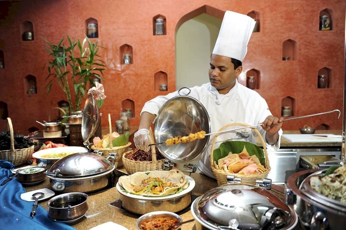 Ein Koch bereitet Speisen auf einem Grill zu, während er in der Küche des Shangri-La Barr Al Jissah Resorts & Spa steht.