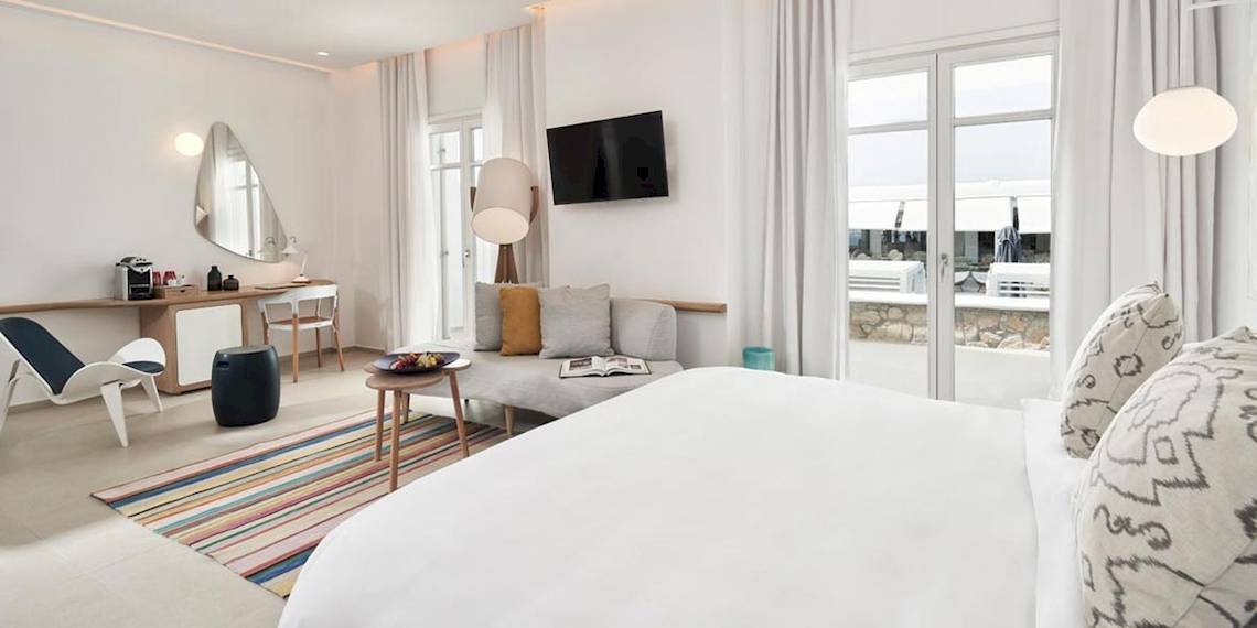 Innenansicht eines Zimmers im Myconian Kyma Hotel mit großem Bett, Couch, Tisch und Fenstern.