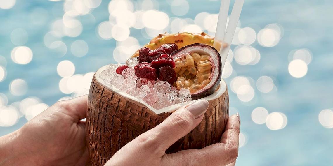 Cocktail im Kokosnuss-Becher mit Früchten und Eis, gehalten von Händen neben einem Pool im Myconian Kyma Hotel