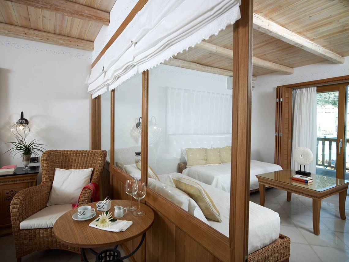 Innenansicht eines Schlafzimmers im Mitsis Blue Domes Resort & Spa mit einem Bett, einem Tisch, einem Stuhl und einem Glasfenster.