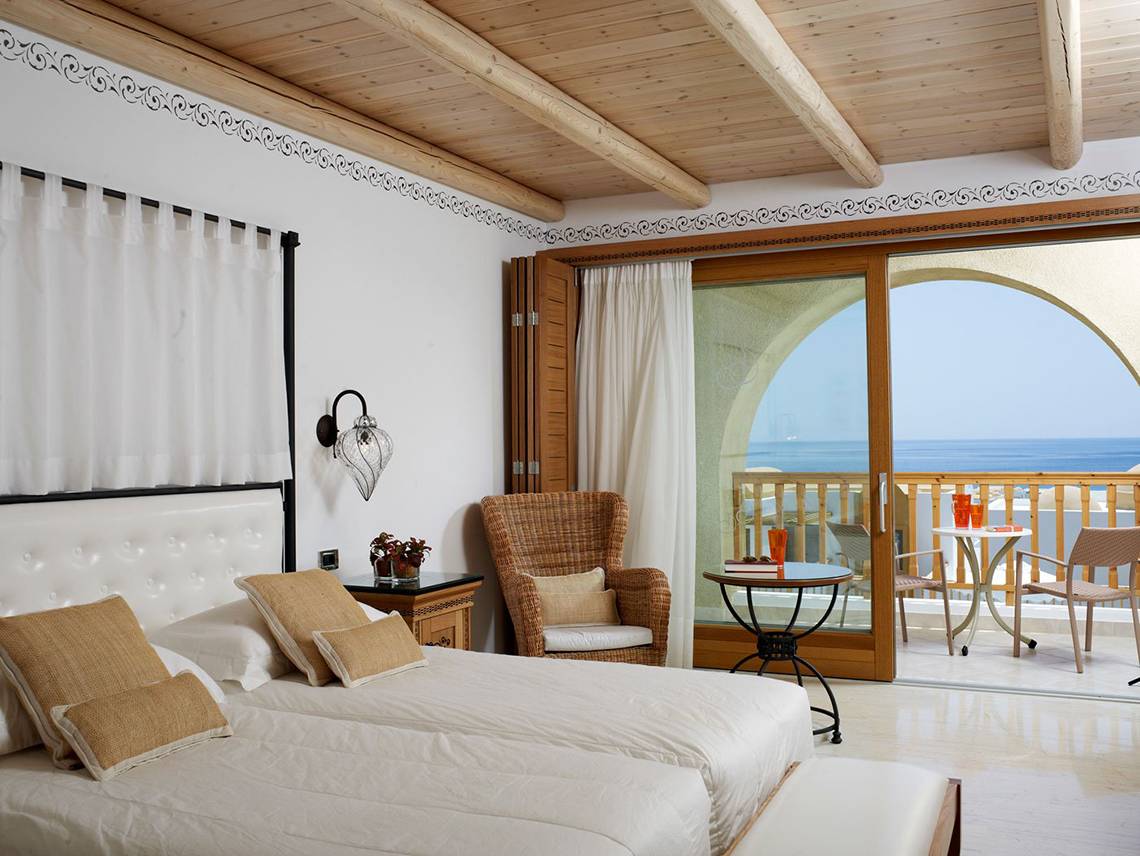 Schlafzimmer im Mitsis Blue Domes Resort & Spa mit Meerblick, inklusive Doppelbett, Sessel, Nachttisch und Balkon mit Tisch und Stühlen.