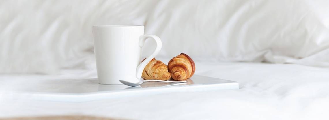 Ein weißer Kaffeebecher, zwei Croissants und ein Löffel auf einem Tablett auf einem Bett.