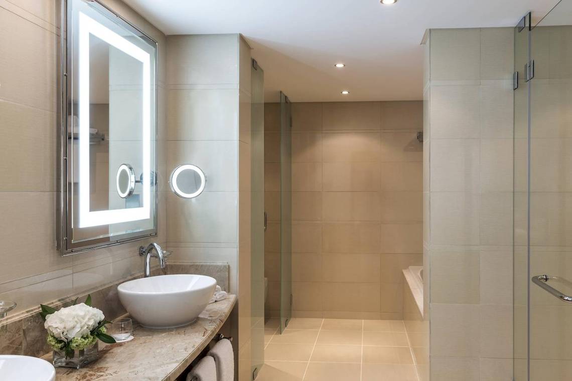 Badezimmer des Sheraton Oman Hotels mit Waschbecken, Spiegel und begehbarer Dusche.
