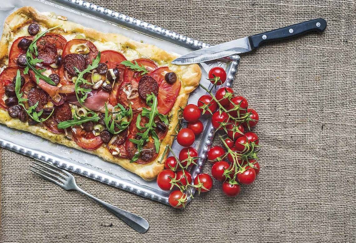Eine Pizze mit Tomaten und Rucola auf einem Tablett neben Tomaten und einem Messer