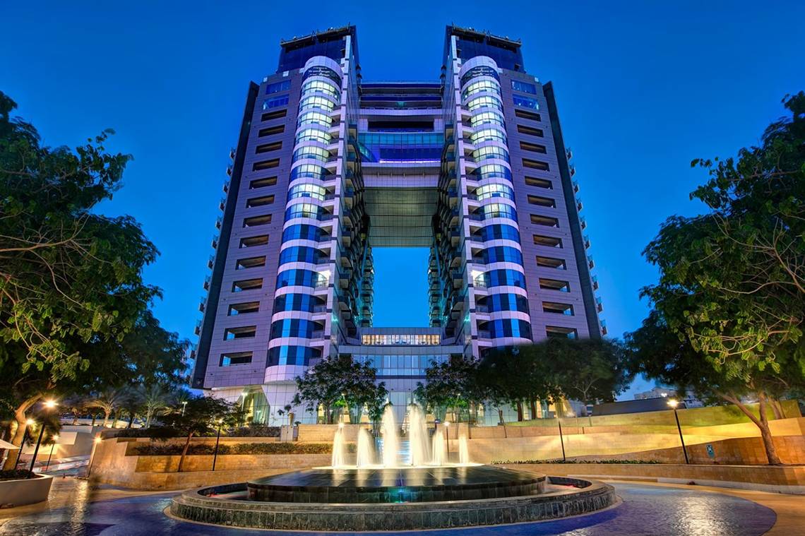 Ein modernes Gebäude namens Dukes The Palm, ein Royal Hideaway Hotel, steht bei Nacht mit blauer Beleuchtung und einem Brunnen davor.