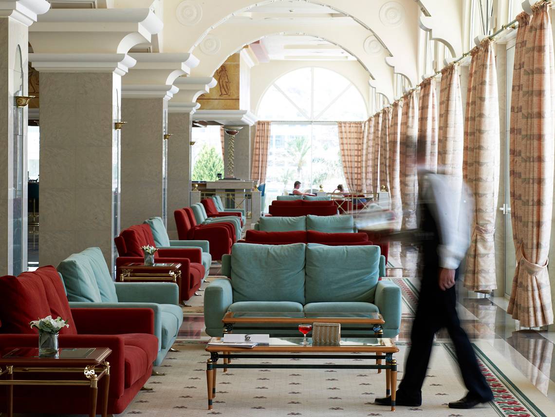 Loungebereich eines Hotels mit roten und blauen Sofas, einem Glastisch und einem vorbeigehenden Mitarbeiter.