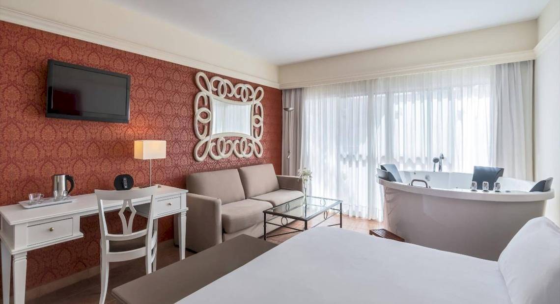 Luxuriöses Zimmer im Barcelo Corralejo Bay Hotel mit rotem Wanddesign, Fernseher, Schreibtisch, Couch, Badewanne und großer Fenster mit Vorhängen.