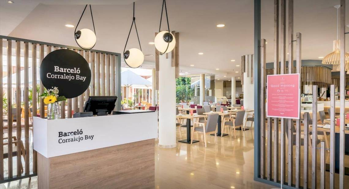 Innenansicht der Rezeption des Barcelo Corralejo Bay Hotels mit Tisch, Stühlen und Schildern.