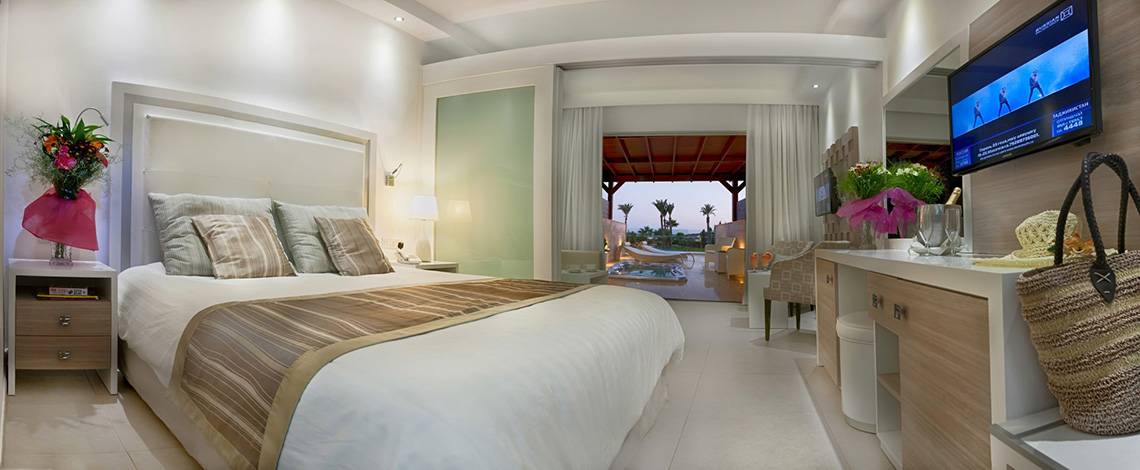 Innenansicht eines Schlafzimmers im Asterias Beach Hotel mit großem Bett, Fernseher, Tisch und Balkon.