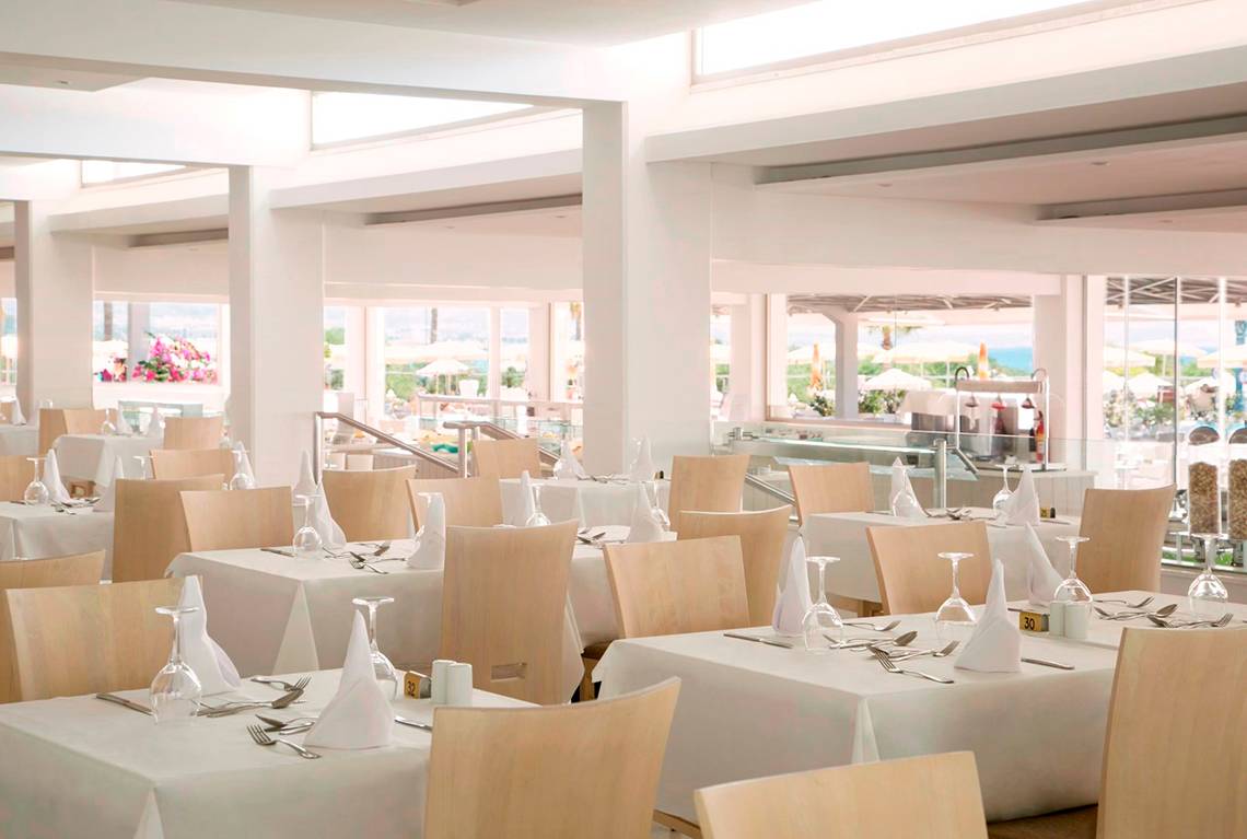 Innenansicht des Restaurants im Asterias Beach Hotel mit gedeckten Tischen und Blick auf das Meer.