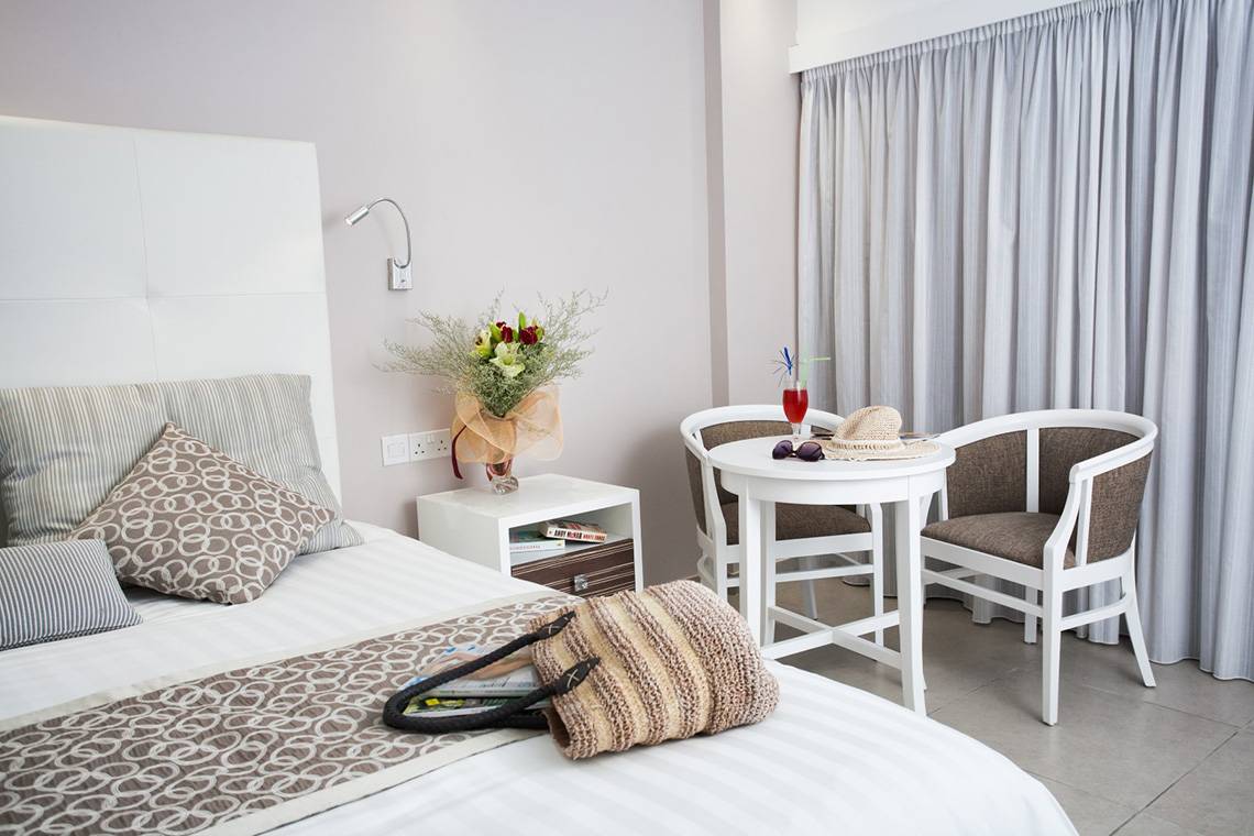 Hotelzimmer im Asterias Beach mit Bett, Tisch, Stühlen, Blumenvase und Tasche.