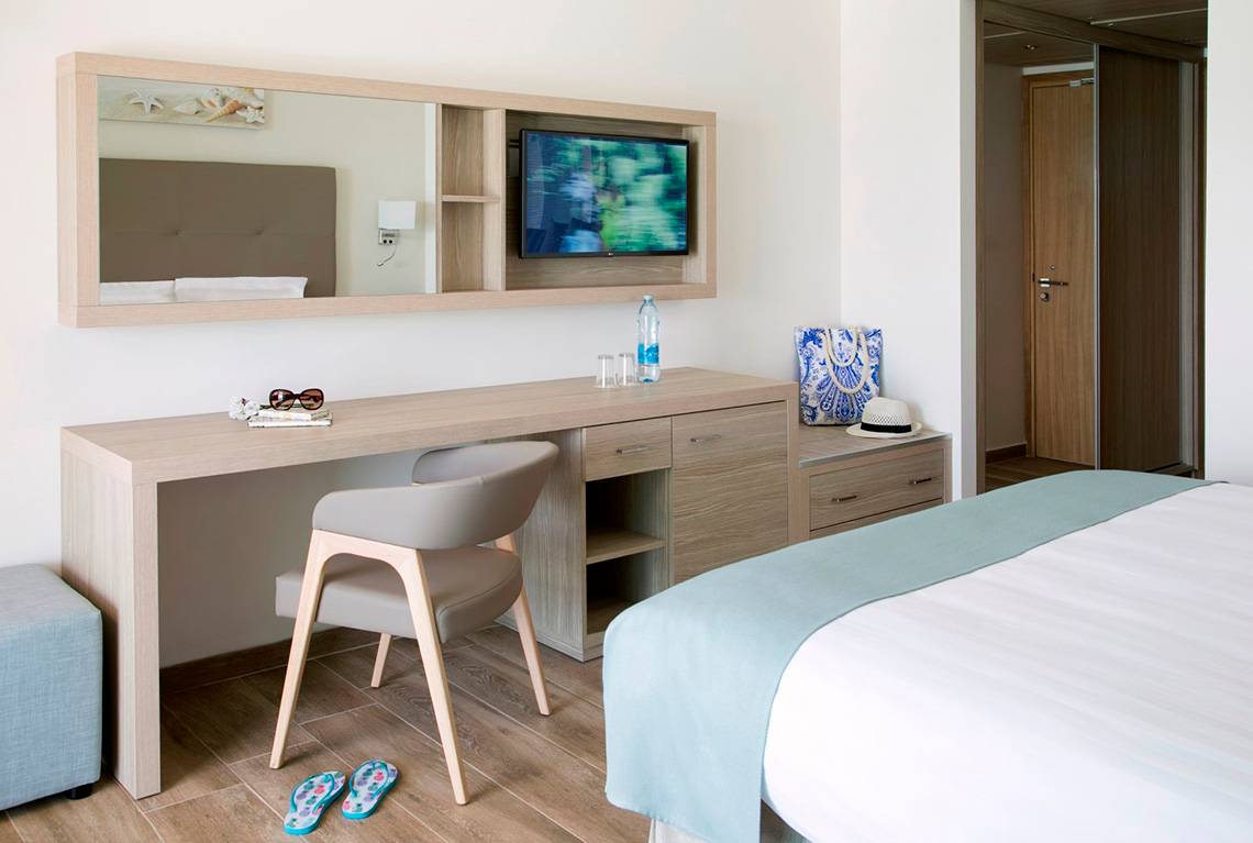 Innenansicht eines Zimmers im Asterias Beach Hotel mit Schreibtisch, Stuhl, Fernseher und Bett.