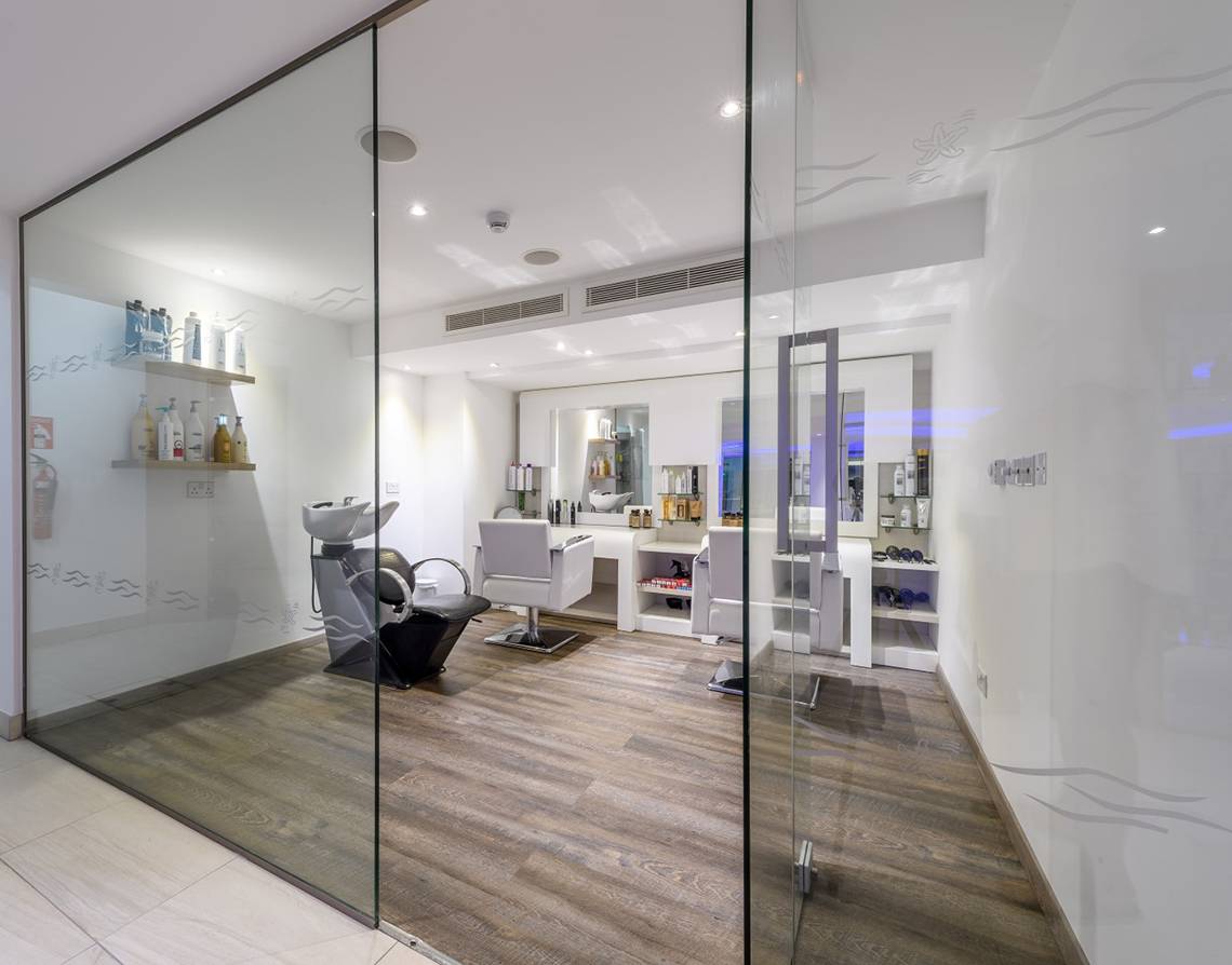 Innenansicht des Friseursalons im Asterias Beach Hotel mit Glaswänden, Holzboden und weißen Möbeln.
