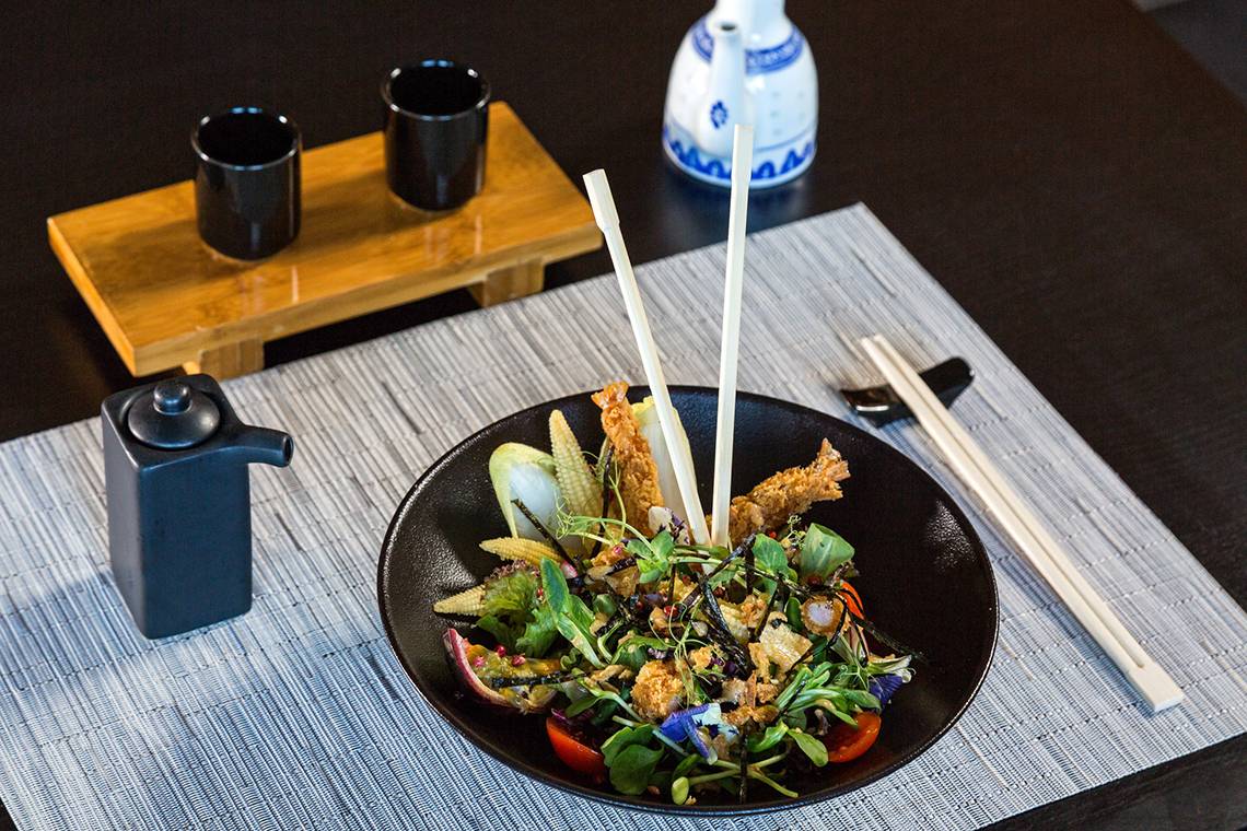 Ein Salat in einem schwarzen Teller mit Besteck auf dem Tisch, umgeben von Utensilien.